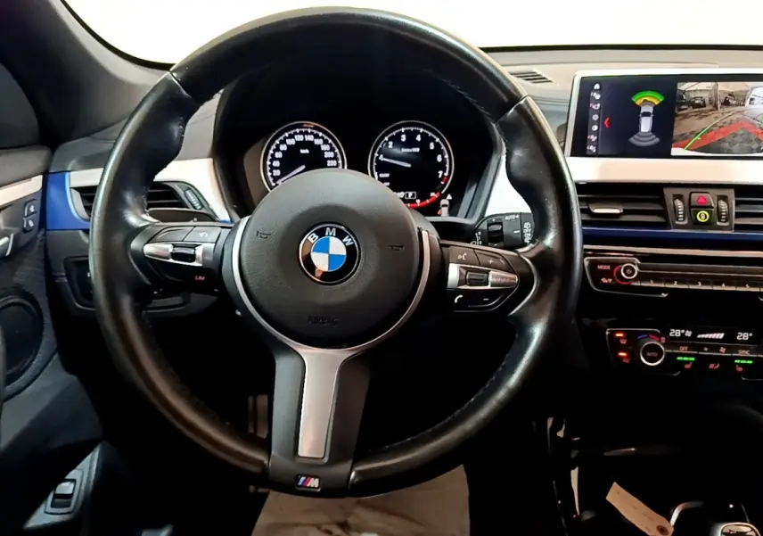 Vue intérieure centrée sur le volant cuir BMW M avec tableau de bord digital et écran tactile 8,8 pouces du BMW X1 blanc.
