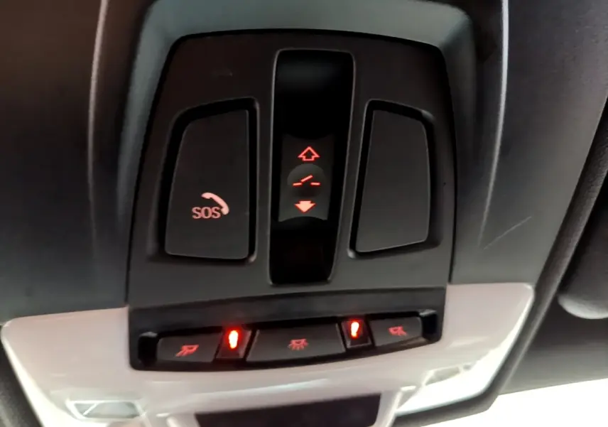 Vue en gros plan du panneau de commandes au plafond du BMW X1 blanc, avec bouton SOS et éclairage intérieur activé.