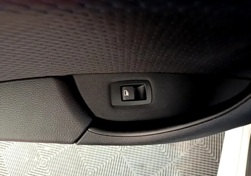 Gros plan sur le bouton de fermeture électrique du coffre, intégré dans la garniture noire de la porte d’un BMW X1 blanc.