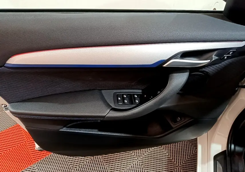 Vue intérieure côté gauche de la porte avant d’une BMW X1 blanche, avec insert décoratif tricolore et commandes de vitres.