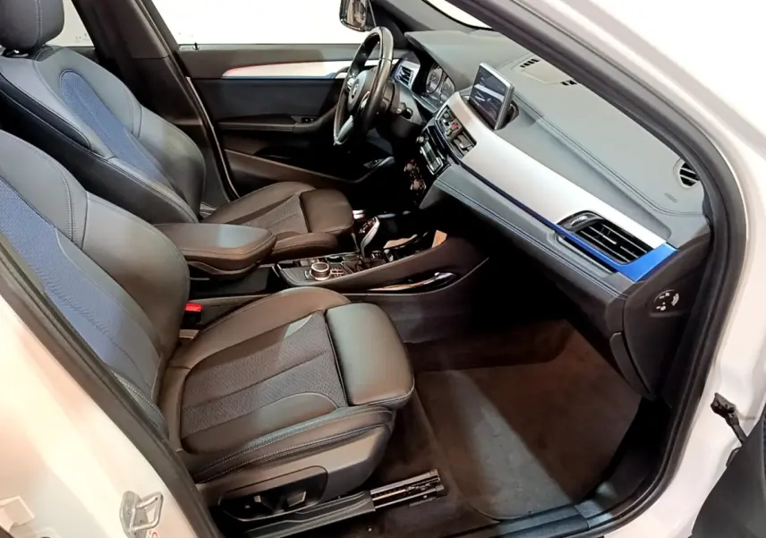 Vue intérieure côté conducteur d’un BMW X1 blanc 2022, sièges cuir noir avec inserts tissu bleu et tableau de bord moderne.