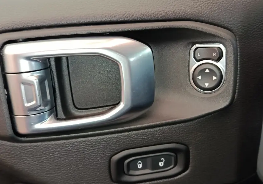 Gros plan sur la poignée intérieure argentée et les commandes de réglage des rétroviseurs du côté conducteur d’un Jeep Wrangler gris foncé.
