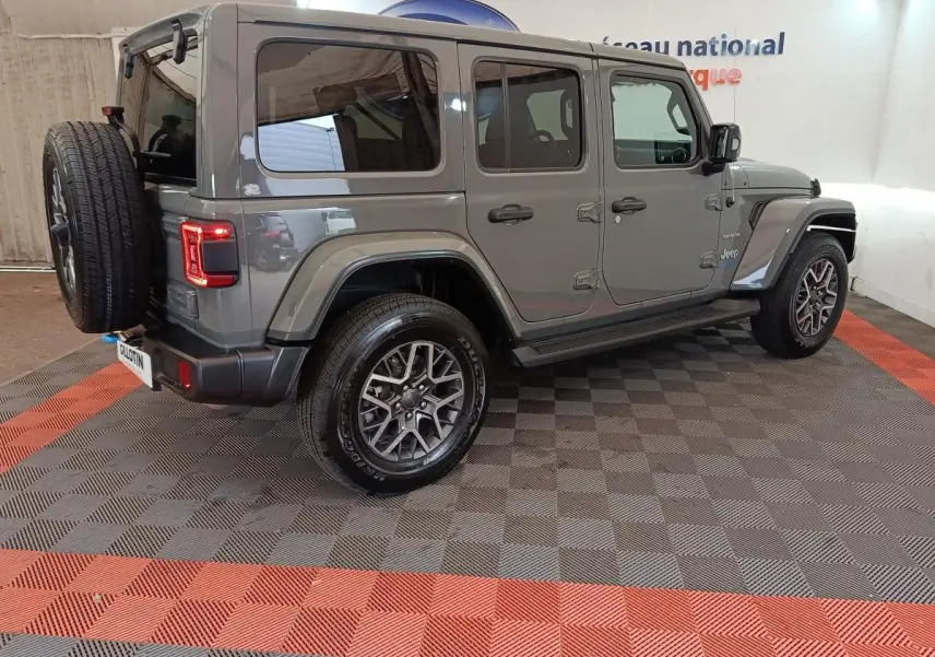 Jeep Wrangler gris foncé vue 3/4 arrière droit, avec roue de secours montée à l'arrière et feux LED allumés.