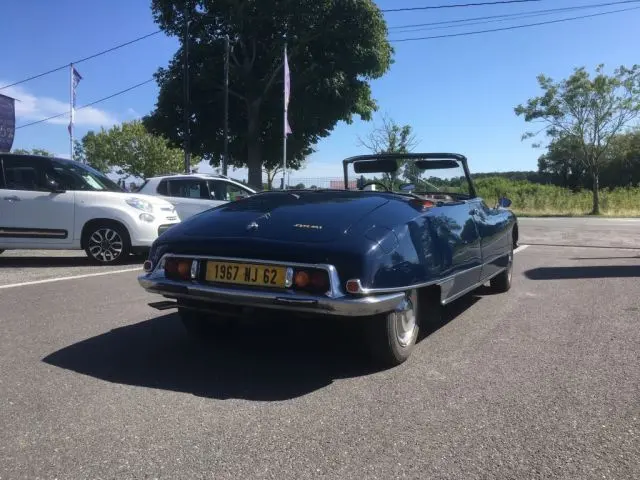 Vue 3/4 arrière d'une Citroën DS3 DS21 Cabriolet Chapron bleu marine de 1967, capote ouverte, sur parking ensoleillé.