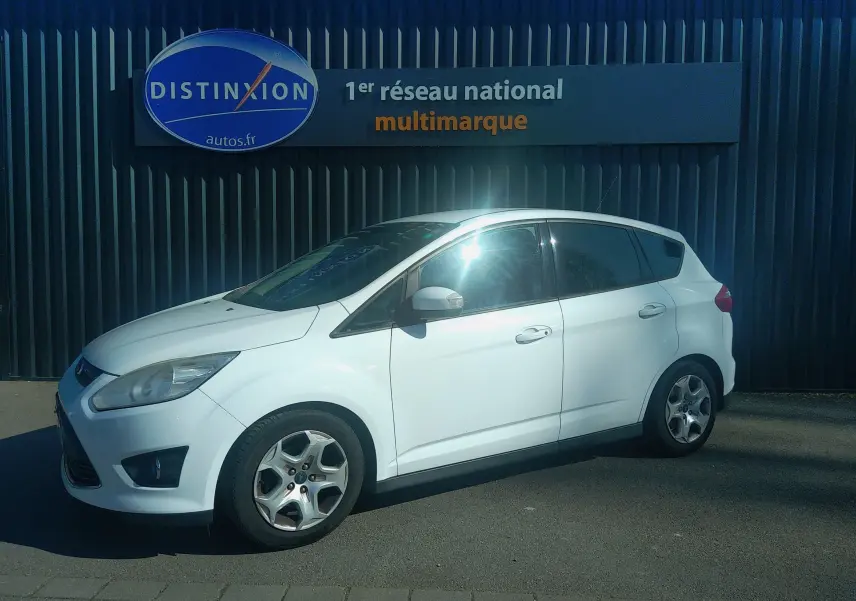 Ford C-MAX blanc en vue de profil côté gauche, garée devant un mur avec panneau publicitaire multimarque.