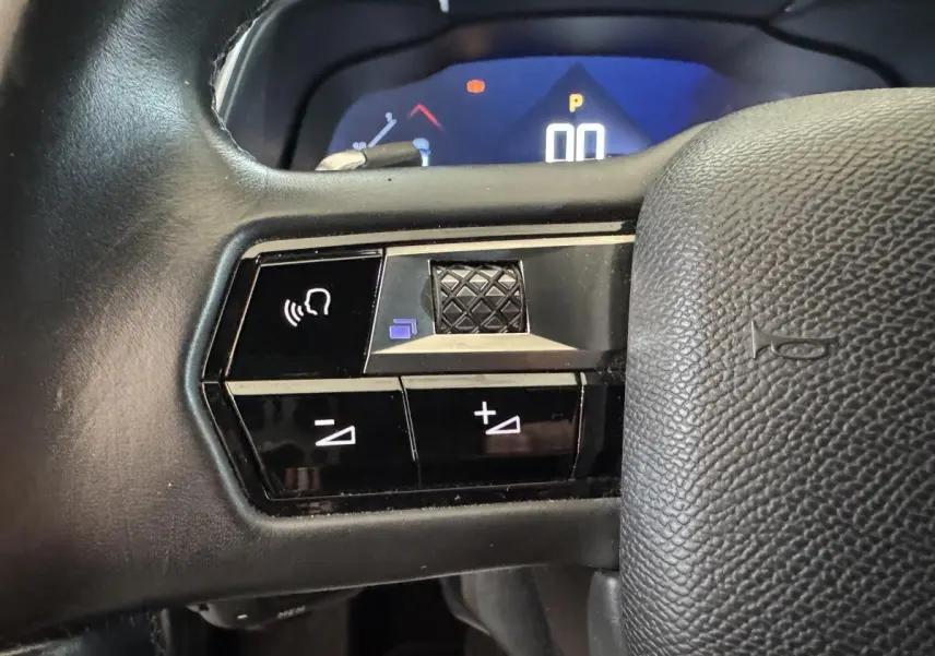 Gros plan sur les commandes vocales et volume au volant cuir noir du DS7 Crossback gris foncé, tableau de bord numérique en arrière-plan.