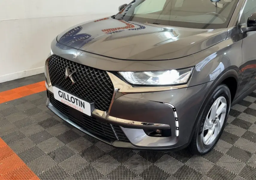 Vue 3/4 avant droit du DS7 Crossback gris foncé avec calandre distinctive et feux LED allumés en intérieur showroom.
