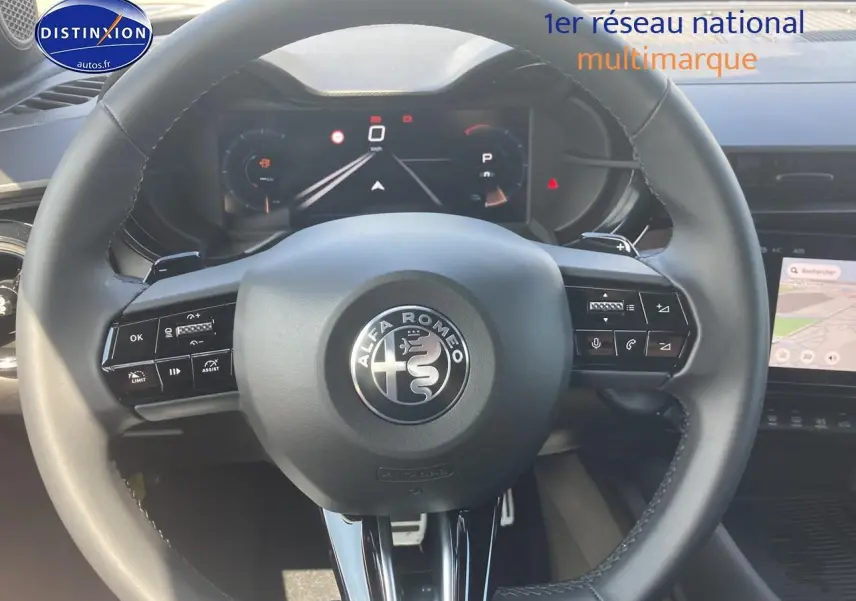 Vue rapprochée du volant noir de l'Alfa Romeo Junior 1.2 hybride 2026 avec tableau de bord digital et écran tactile visible.