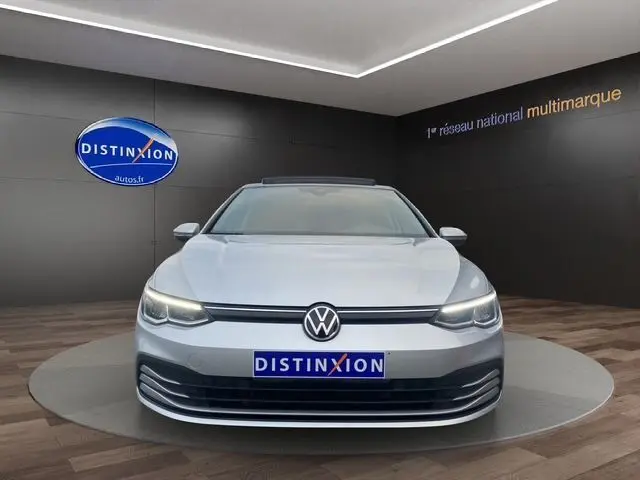 Vue frontale d'une Volkswagen Golf VIII gris argent avec toit panoramique ouvrant et vitres surteintées dans un showroom.