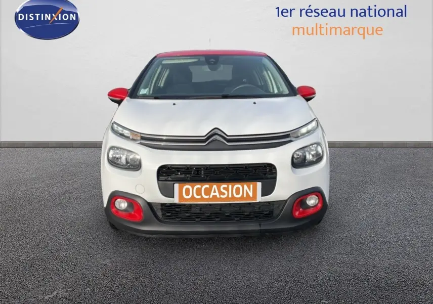 Vue frontale d'une Citroën C3 blanche avec toit et détails rouges, sur fond neutre avec panneau publicitaire.