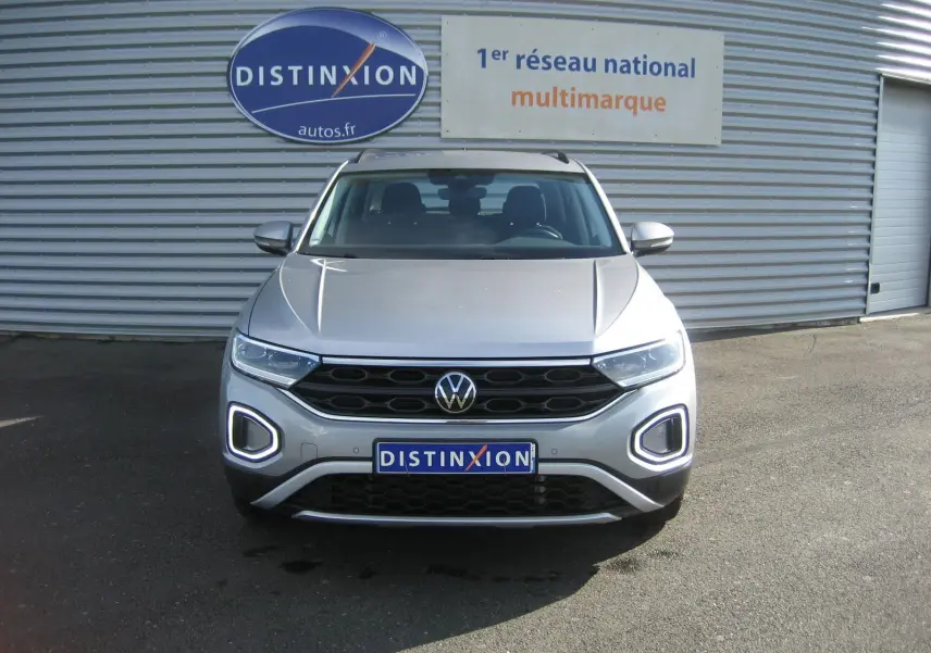 Vue frontale d'un Volkswagen T-Roc gris clair 2023 avec feux LED et calandre noire, devant un mur métallique.