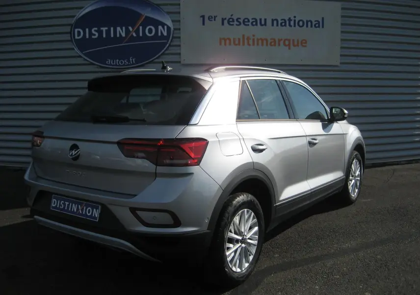 Vue 3/4 arrière droite d’un Volkswagen T-Roc gris clair 2023 garé devant un mur métallique avec logo Distinxion.