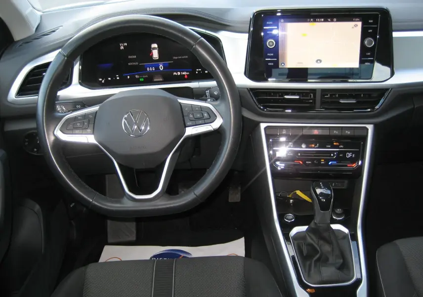 Vue intérieure frontale du volant et tableau de bord du Volkswagen T-Roc gris clair avec écran tactile et levier de boîte automatique.