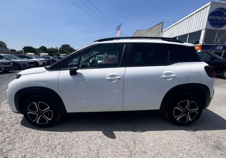 Profil côté gauche d'un Citroën C3 Aircross blanc banquise 2023 avec toit blanc opale et barres de toit noires brillantes.