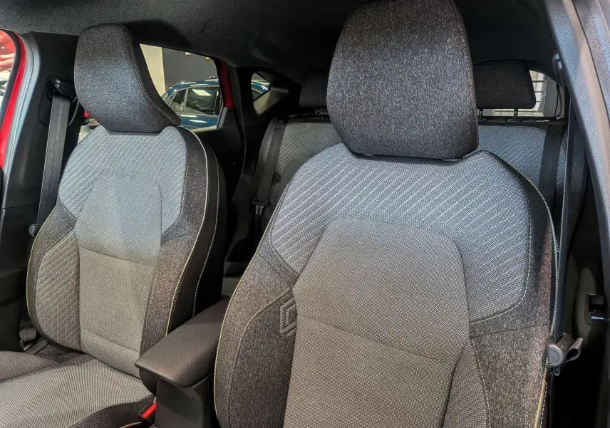 Gros plan sur les sièges avant en tissu gris du Renault Captur E-TECH 2025, avec portières rouges visibles en arrière-plan.
