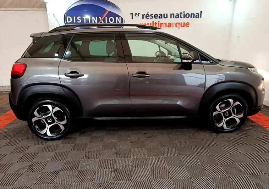 Vue de profil côté gauche d’un Citroën C3 Aircross gris foncé avec barres de toit noires en intérieur showroom.