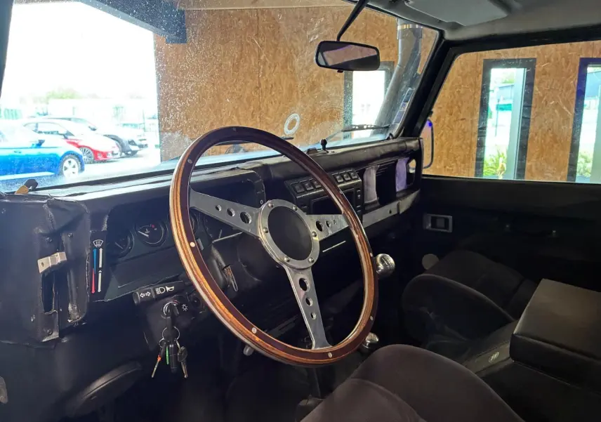Intérieur du Land Rover Defender 110 vert de 2005, vue côté conducteur, volant bois vintage et tableau de bord simple.