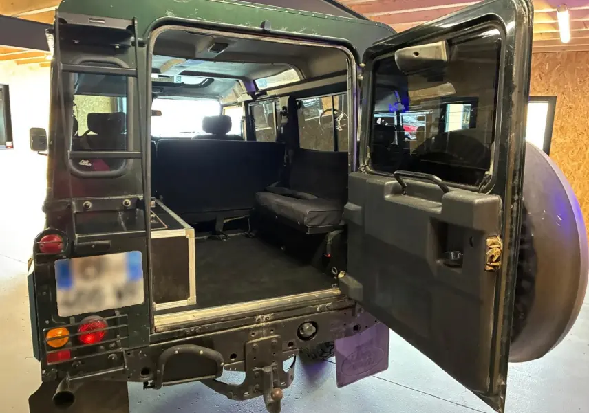 Vue arrière ouverte du Land Rover Defender 110 vert 2005, montrant l'intérieur spacieux et les sièges latéraux.