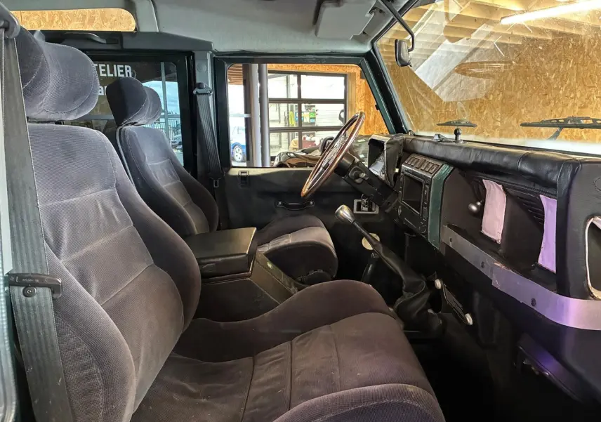 Vue intérieure côté conducteur du Land Rover Defender 110 2005, montrant les sièges noirs et le tableau de bord avec volant bois.