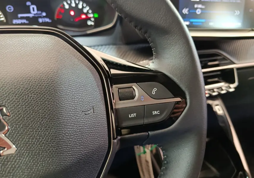 Gros plan sur le volant cuir multifonctions de la Peugeot 208 blanche, avec commandes audio et tableau de bord en arrière-plan.