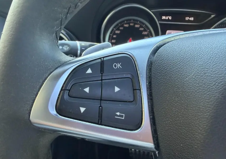 Gros plan sur les commandes au volant côté gauche du Mercedes GLA 220d blanc, tableau de bord en arrière-plan.
