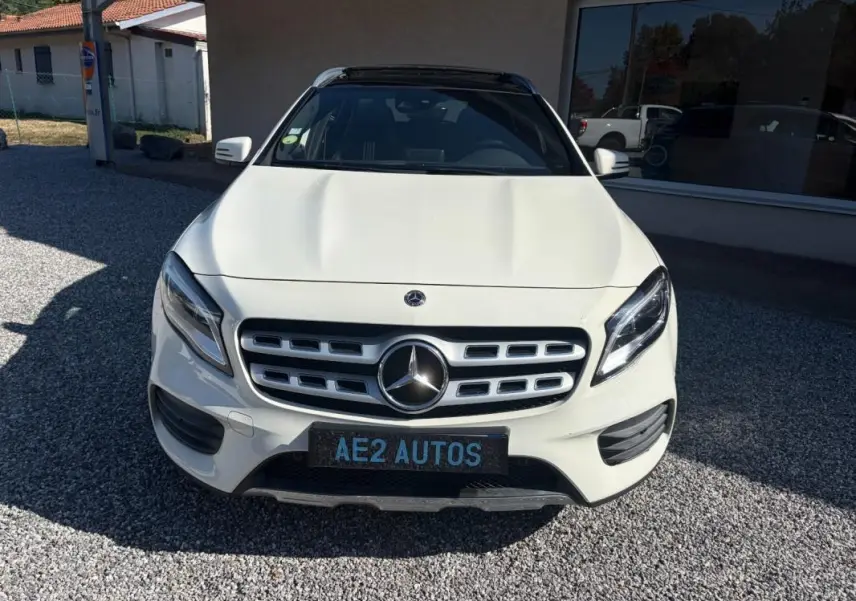 Vue avant d'un Mercedes GLA 220d blanc avec calandre chromée et toit panoramique noir, stationné sur gravier devant un bâtiment.