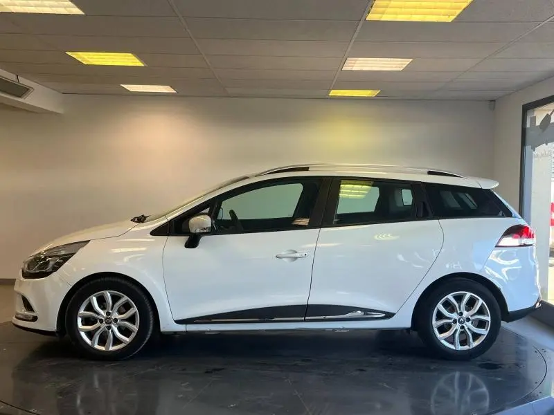 Profil droit d'une Renault Clio Estate blanc glacier 2019 avec barres de toit et jantes alliage visibles en intérieur showroom.