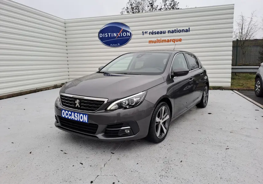 Peugeot 308 gris vue 3/4 avant droit avec jantes alliage et calandre chromée sur parking extérieur.