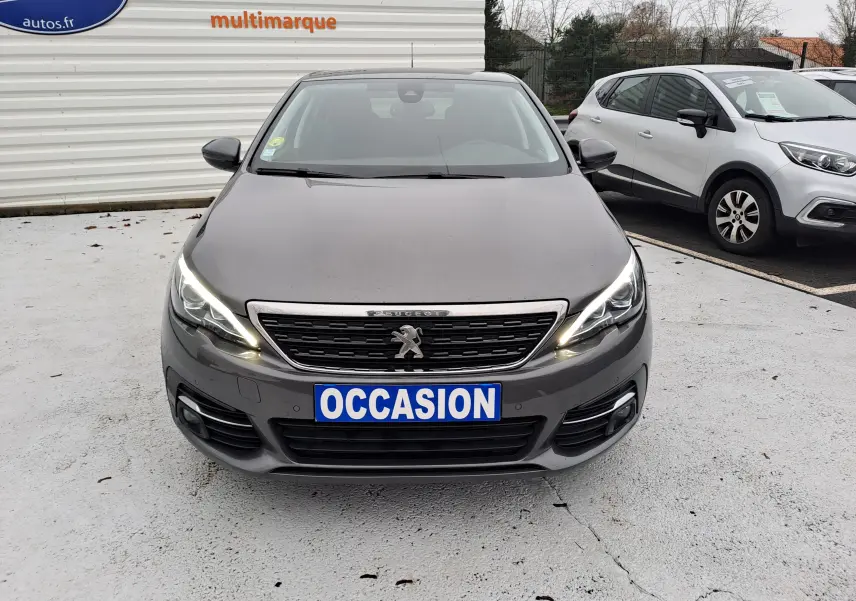 Peugeot 308 gris vue de face avec phares allumés et plaque "OCCASION" sur un parking extérieur.