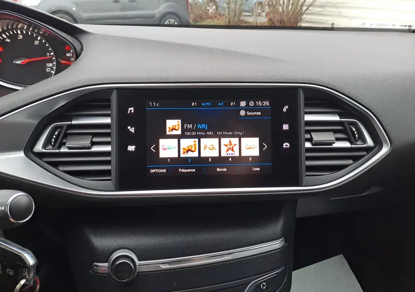 Tableau de bord noir de Peugeot 308 gris 2019 avec écran tactile central affichant les radios FM, vue intérieure frontale.