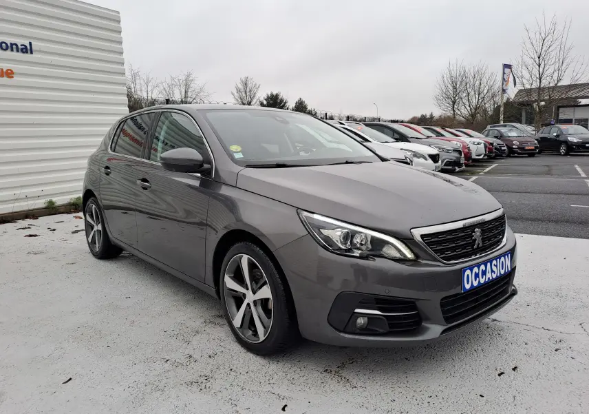 Peugeot 308 gris en 3/4 avant droit avec jantes alliage et plaque "OCCASION" visible sur un parking.