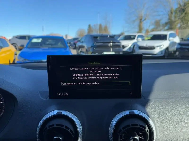 Vue intérieure du tableau de bord de l'Audi Q2 30 TDI S line 2024 avec écran central et aérateurs ronds, extérieur visible au loin.