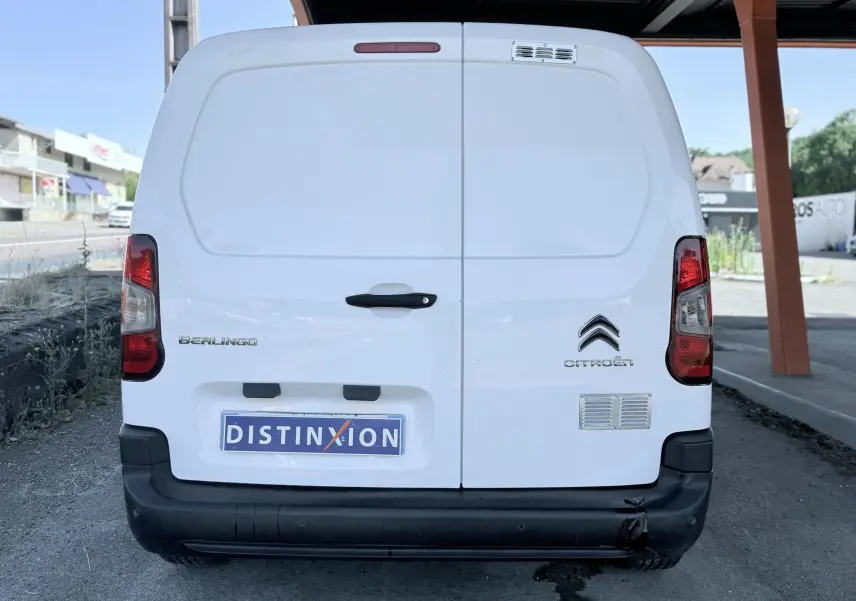 Vue arrière d’un utilitaire blanc Citroën Berlingo avec logo et plaques visibles, sous un abri métallique.