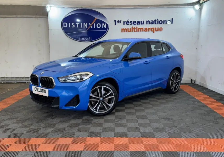BMW X2 xDrive25e M Sport bleu en 3/4 avant droit dans un showroom avec sol gris et bandes orange.