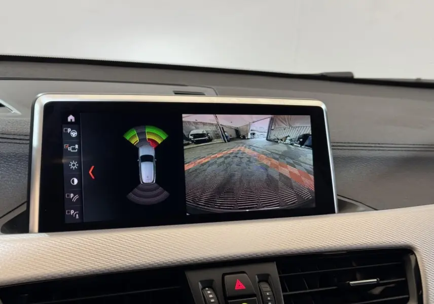 Écran central de BMW X2 xDrive25e bleu montrant l’aide au stationnement avec vue caméra arrière et capteurs.
