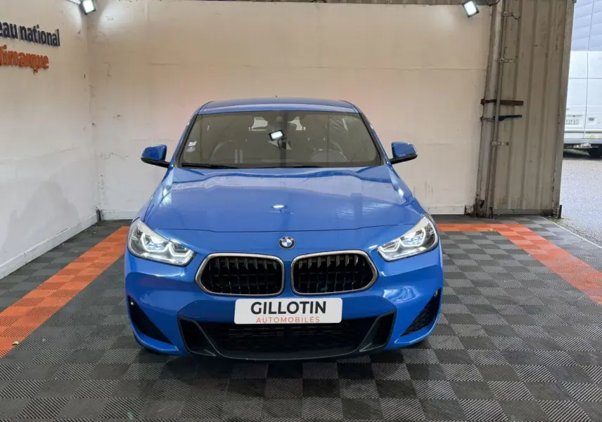 BMW X2 xDrive25e M Sport bleu vue de face avec calandre noire et phares allumés dans un garage.