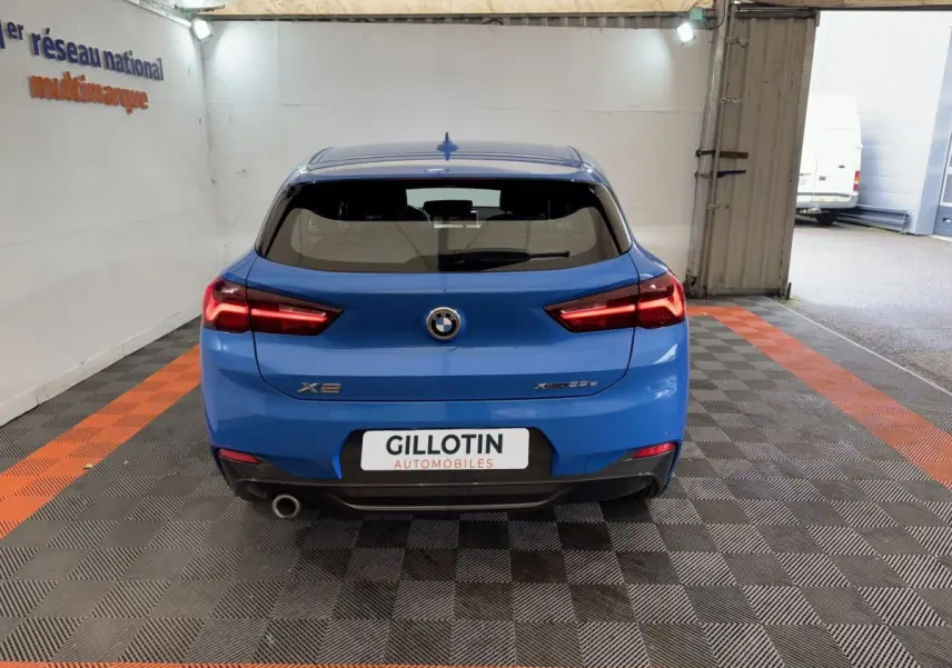 Vue arrière d’un BMW X2 xDrive25e M Sport bleu, avec feux allumés et sortie d’échappement visible côté gauche.