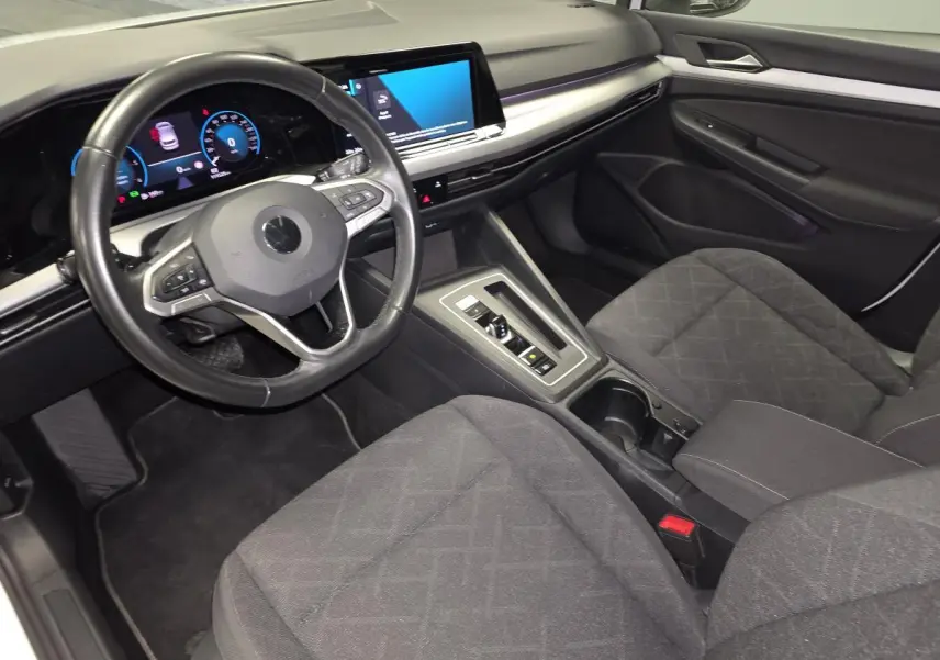 Intérieur de Volkswagen Golf VIII blanc, vue 3/4 avant côté conducteur, tableau de bord et sièges tissu gris visibles.