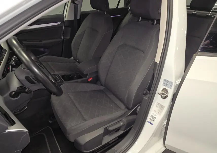 Vue intérieure côté conducteur d'une Volkswagen Golf VIII blanche, sièges avant en tissu gris foncé avec motifs.