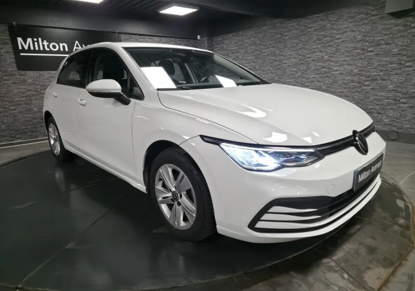 Volkswagen Golf VIII blanche vue en 3/4 avant droit avec phares LED allumés dans un showroom.