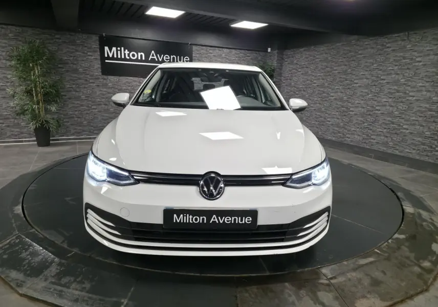 Vue avant d'une Volkswagen Golf VIII blanche avec phares LED allumés dans un showroom moderne.