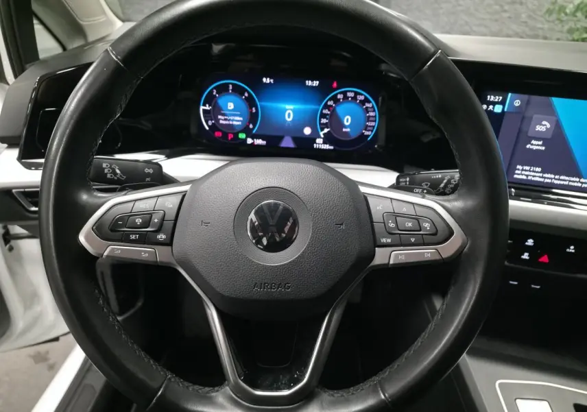 Vue rapprochée du volant noir multifonction et du tableau de bord digital lumineux d'une Volkswagen Golf VIII blanche.