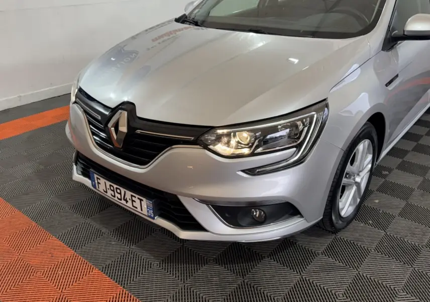 Renault Mégane Business gris clair vue 3/4 avant droit, phare allumé et calandre noire distinctive.