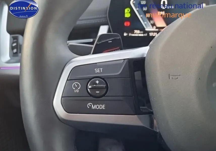 Gros plan sur les commandes de régulateur de vitesse au volant cuir du BMW X1 blanc mineral, tableau de bord numérique en arrière-plan.