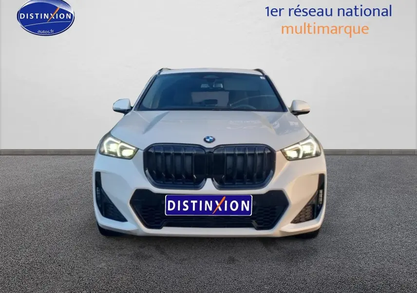 Vue frontale d'un BMW X1 blanc minéral avec calandre noire et phares LED allumés sur fond neutre.