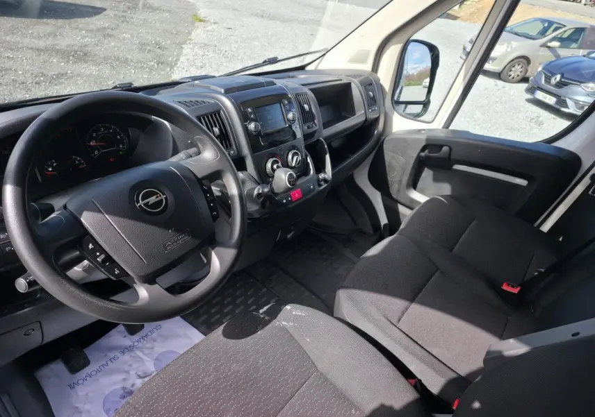 Vue intérieure côté conducteur de l’Opel Movano blanc, montrant le volant, tableau de bord et banquette passagers noire.