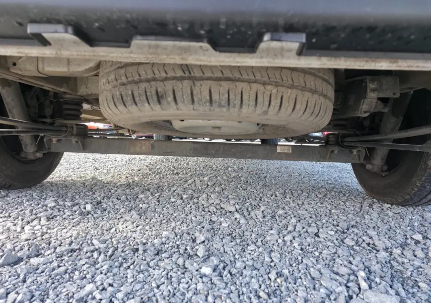 Vue en contre-plongée sous un Opel Movano Fourgon blanc, montrant la roue de secours et le châssis arrière sur sol gravillonné.