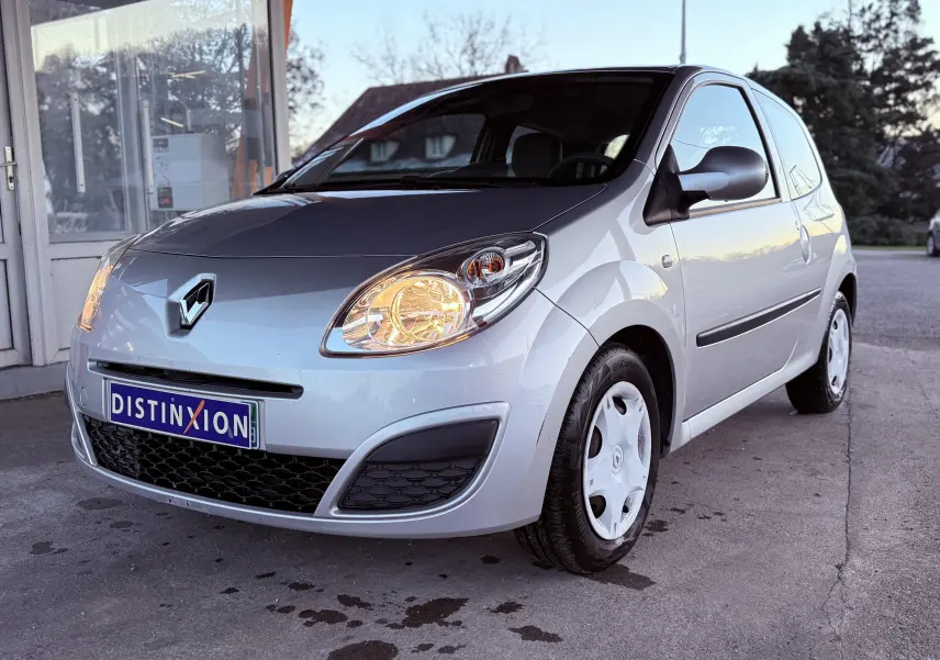 Renault Twingo II gris vue 3/4 avant droit avec phares allumés et jantes tôles blanches.