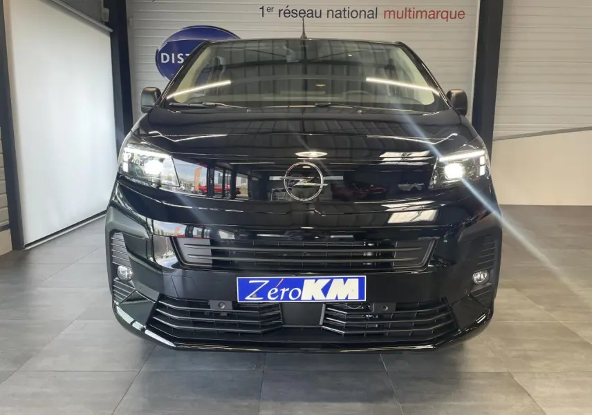 Vue frontale d'un Opel Vivaro noir carbone avec phares allumés et plaque Zéro KM visible en intérieur showroom.