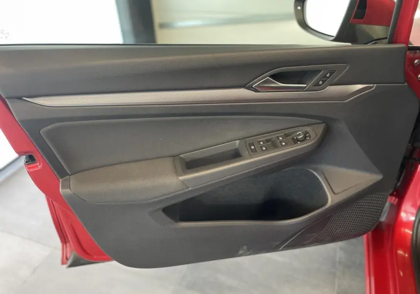 Porte avant gauche ouverte d'une Volkswagen Golf rouge métal 2022, montrant les commandes de vitres et la poignée intérieure chromée.